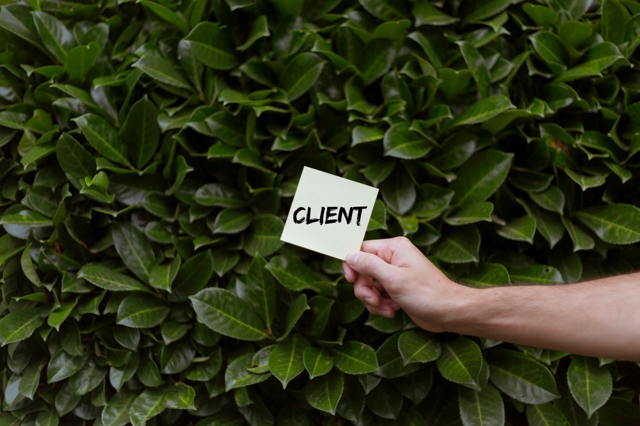 Close-up of a hand holding a note with the word CLIENT in front of a lush green leafy background.